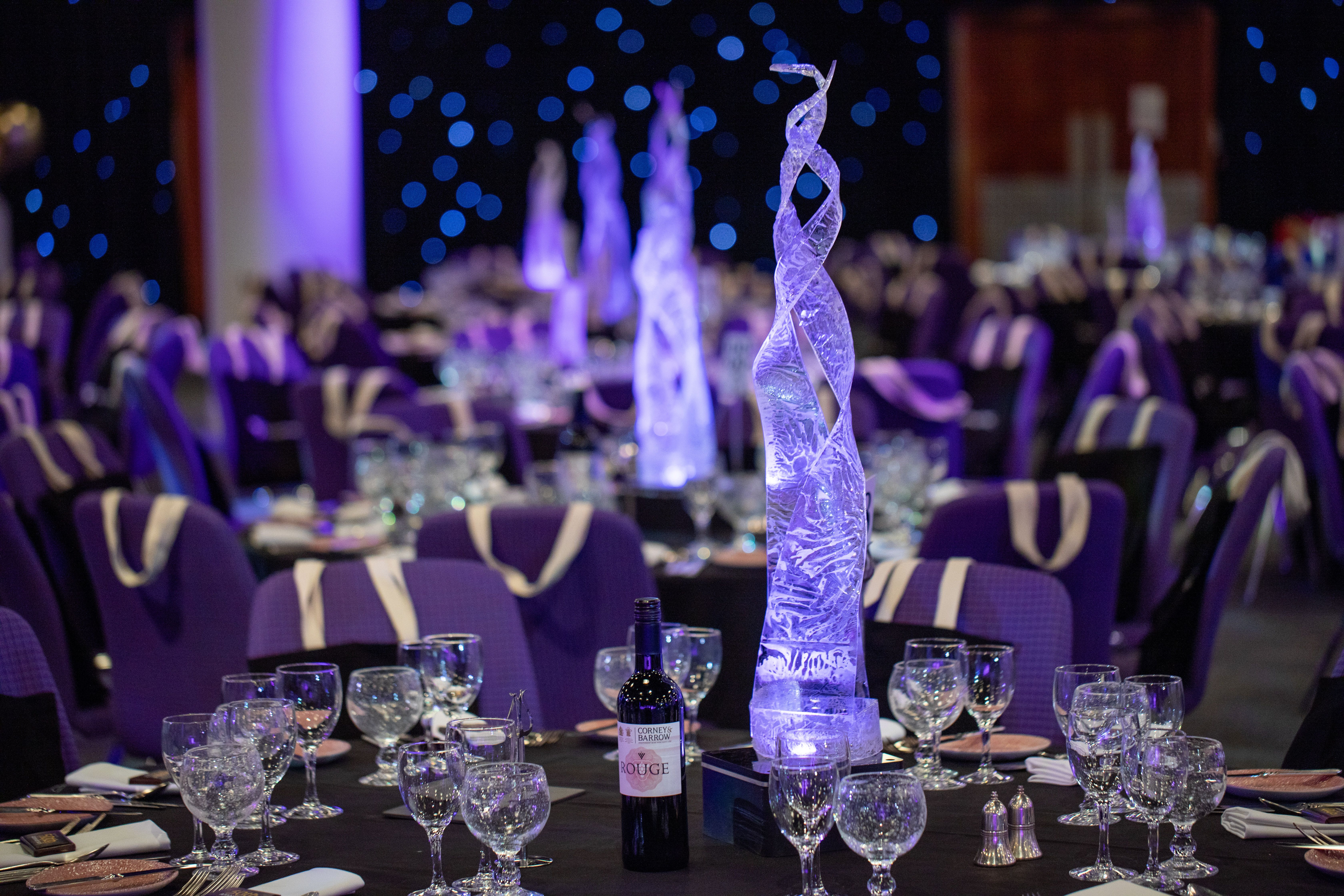 Elegant themed party banquet hall with illuminated ice sculpture centrepieces, black tablecloths and purple chair covers at a corporate evening event by Event Collection