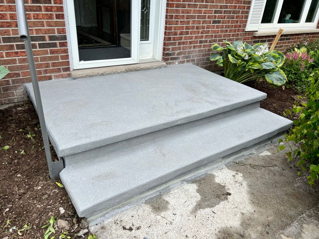 Two-step concrete porch before repair — brick wall entrance with glass door and garden beds