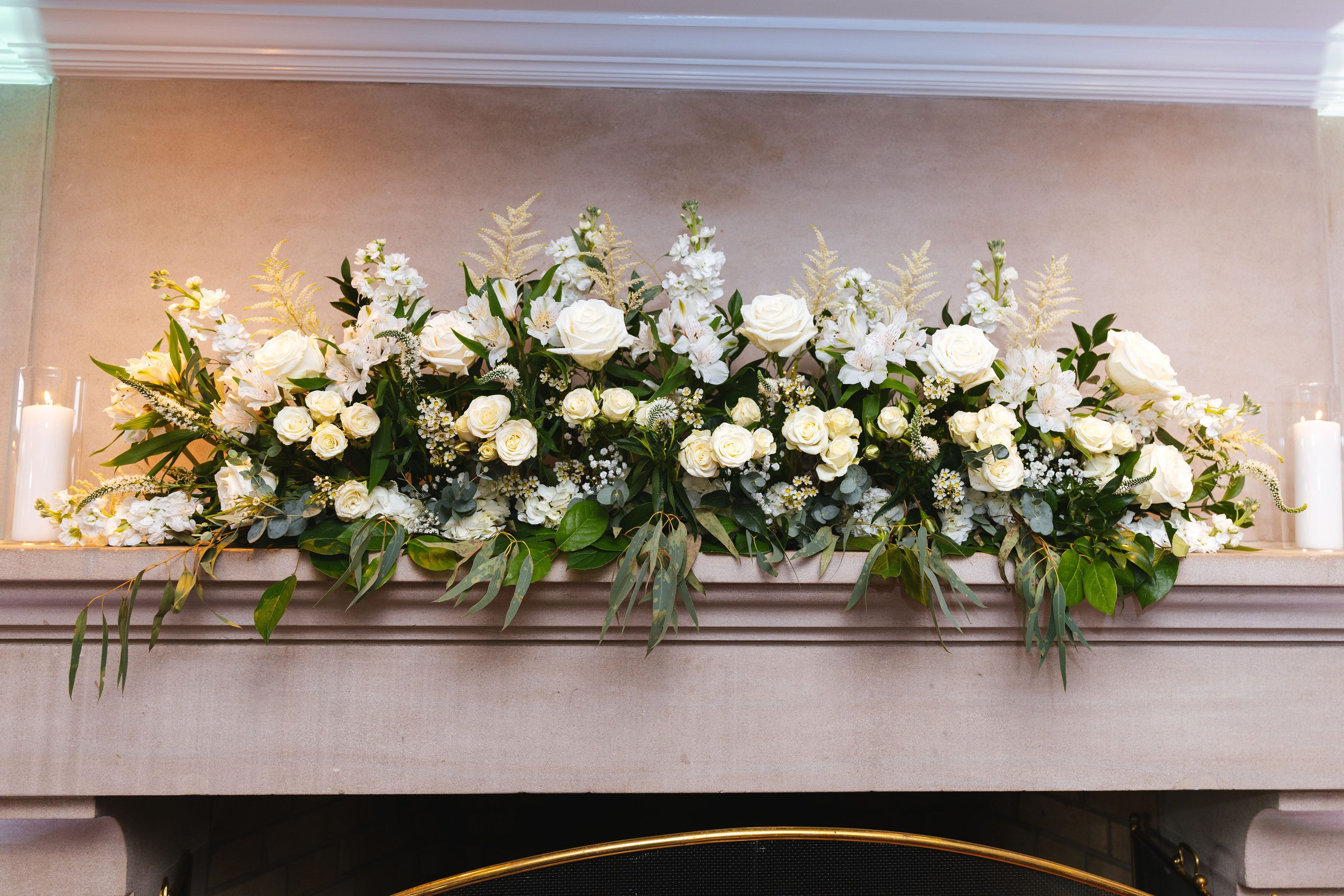 Fireplace mantle adorned with white roses, candles, and cascading greenery