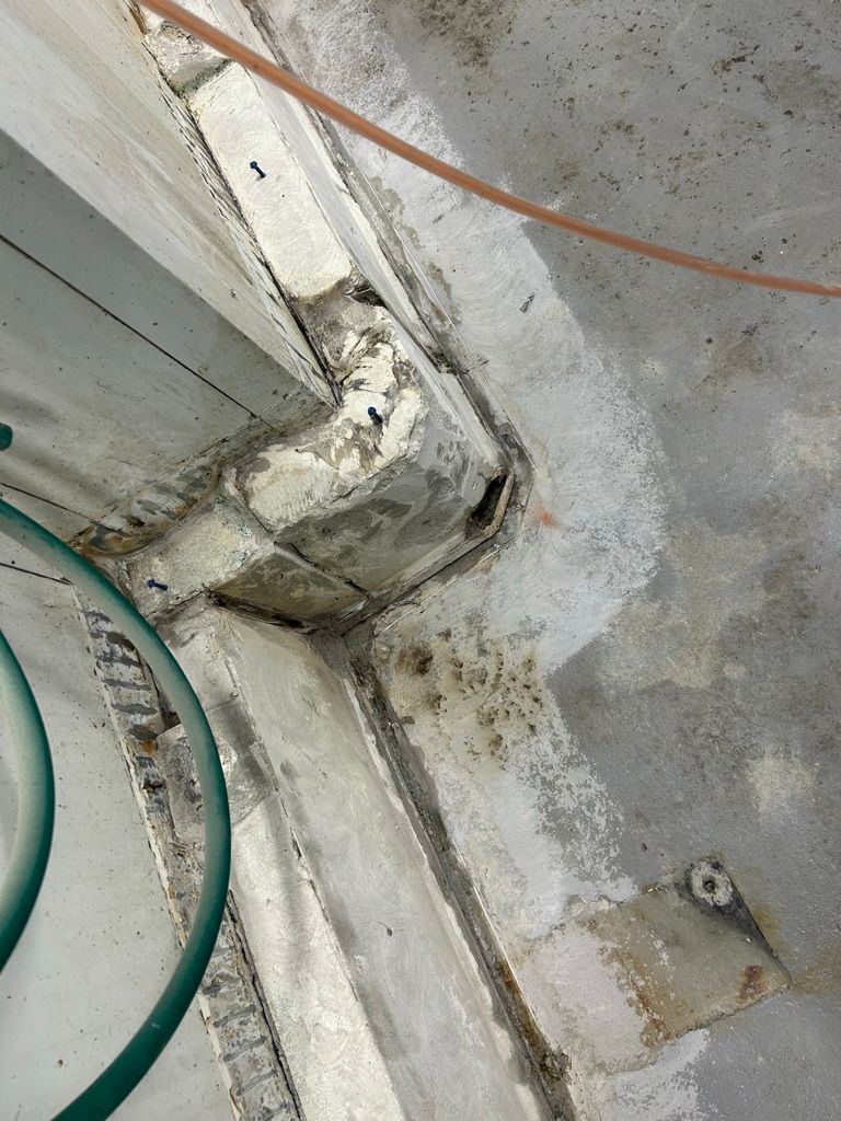 Concrete corner with surface deterioration and anchoring screws alongside green hose before repair