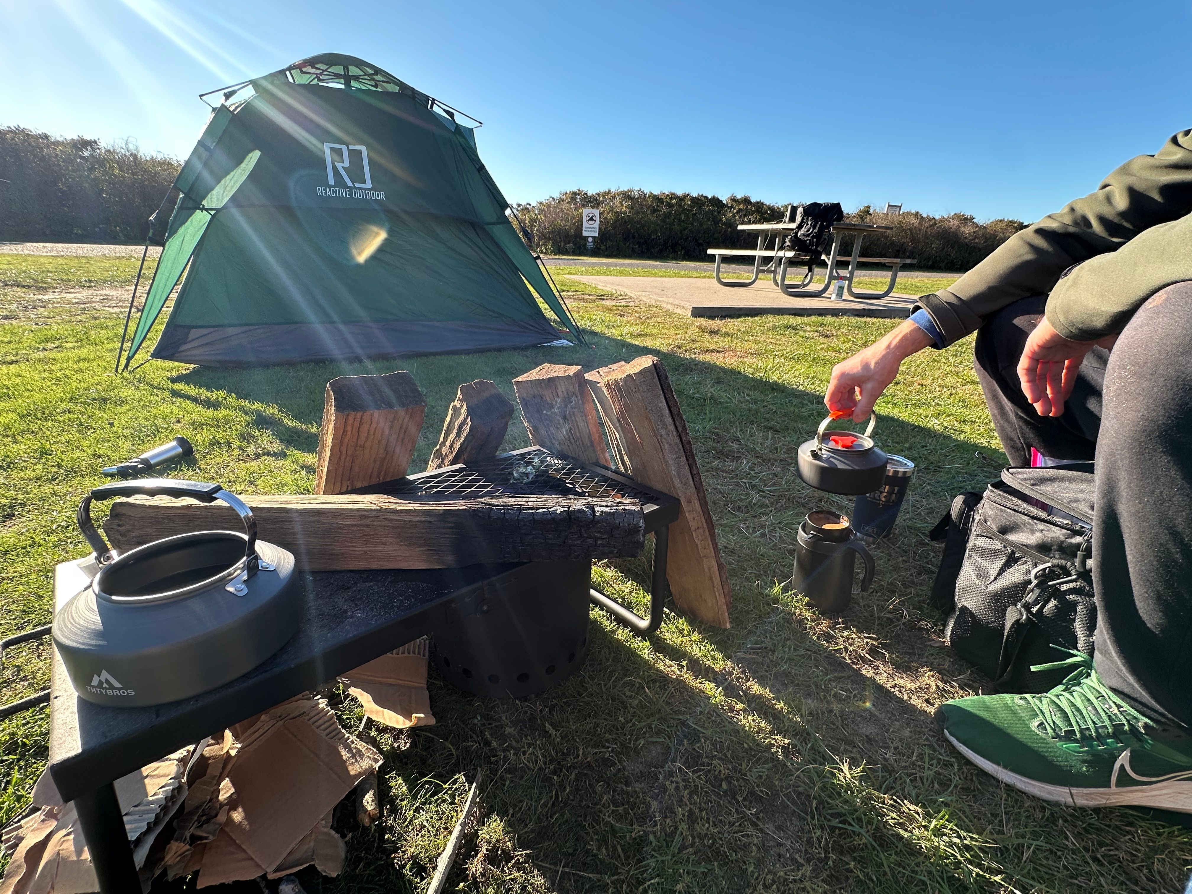 Cooking breakfast at camp — green tent, fire pit, kettle, and morning setup — Hither Hills 2025