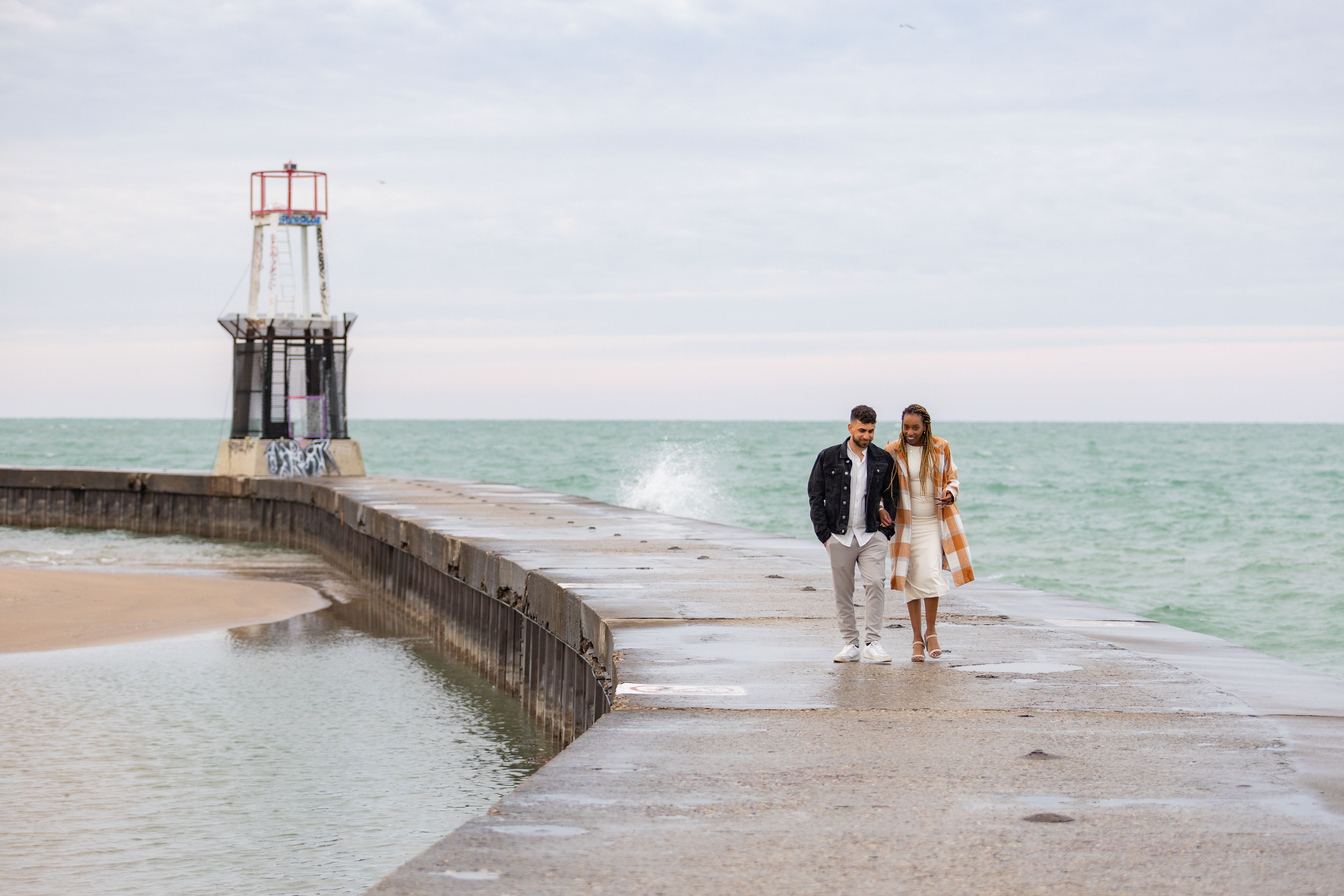 Couple walking on pier toward lighthouse on overcast day