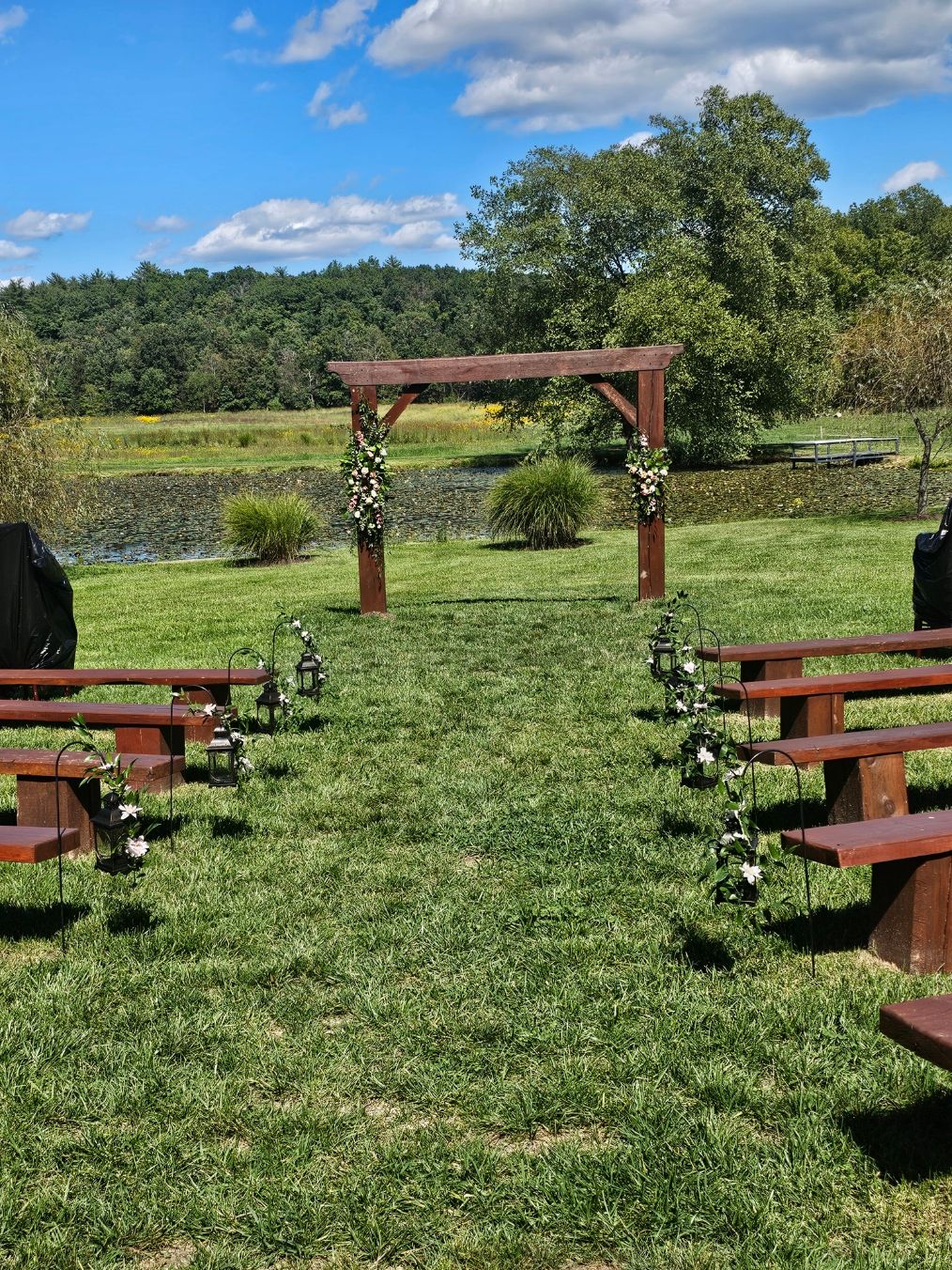 Outdoor ceremony by a pond with wooden arch, lanterns, and floral aisle markers