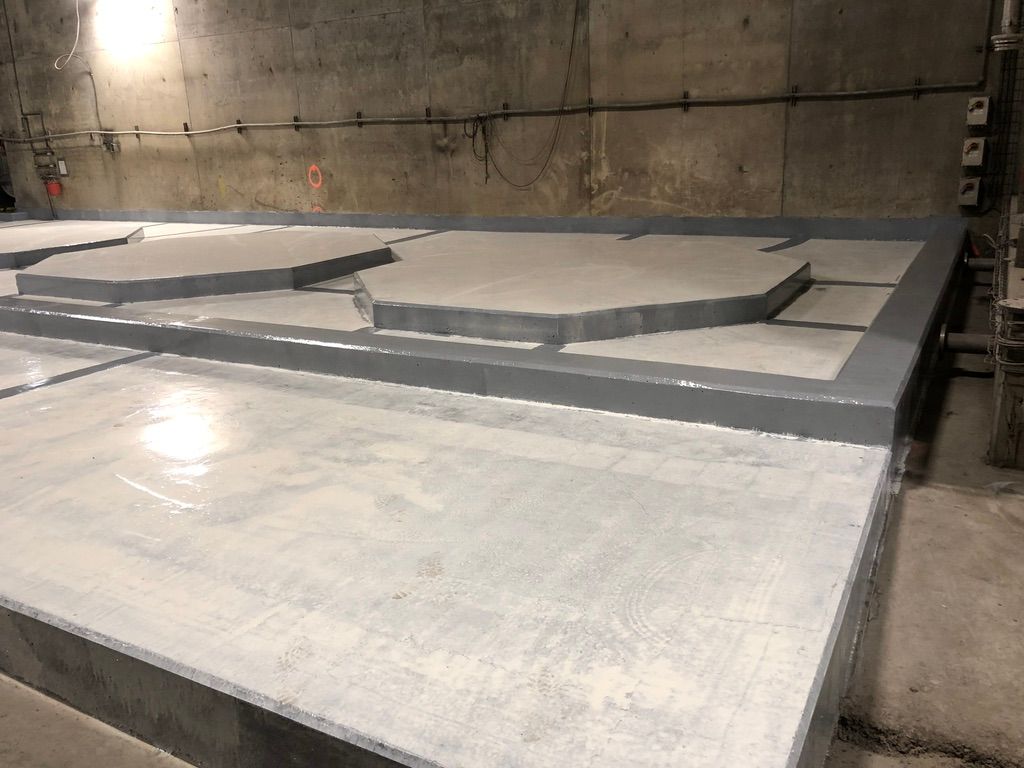 Close-up of water treatment facility floor with raised hexagonal and polygonal platform sections coated in seamless grey epoxy, rough industrial concrete wall with exposed wiring behind