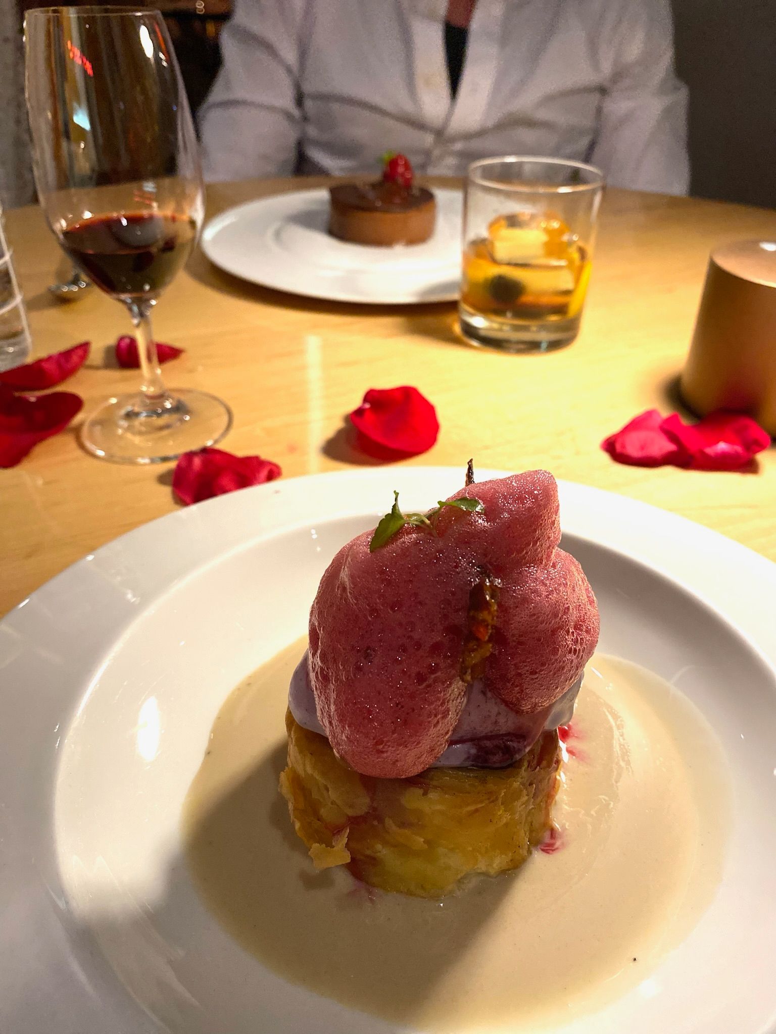 Marmalade tasting menu dessert — layered golden pastry with pale purple cream and pink foam, alongside a glass of red wine and amber cocktail, rose petals on the table