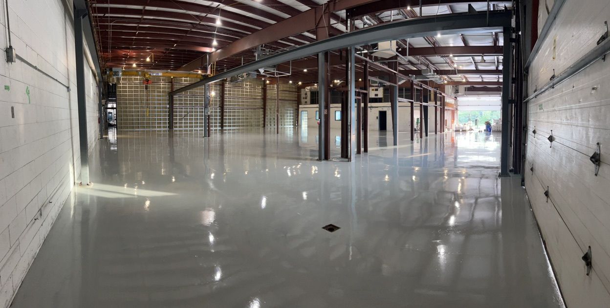 Large empty industrial warehouse with highly polished concrete floor, metal support columns, overhead doors and small enclosed office area