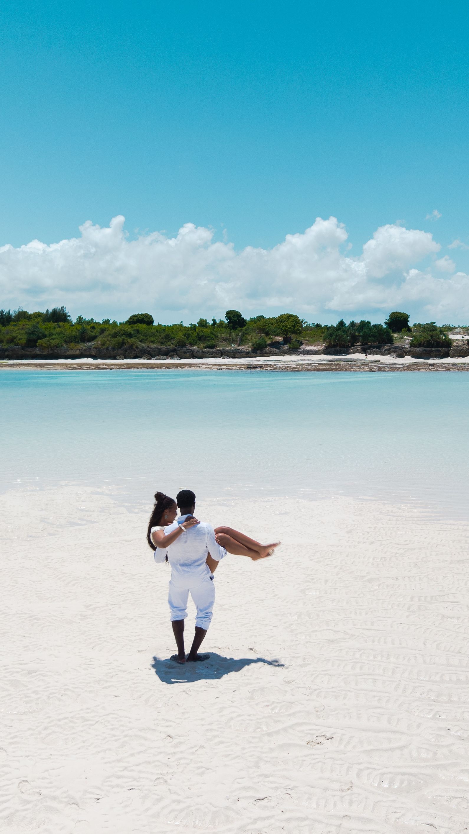 Man carrying woman on white sand beach with turquoise water — drone shot