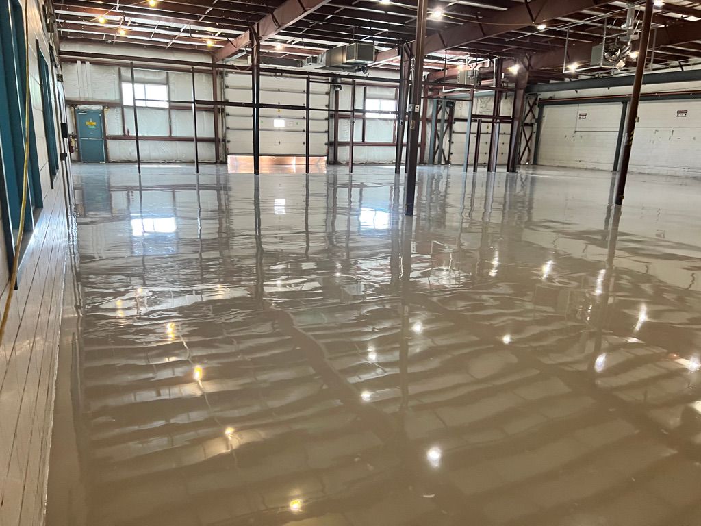 Large empty industrial warehouse with highly reflective smooth floor, exposed metal beams, windows and garage doors