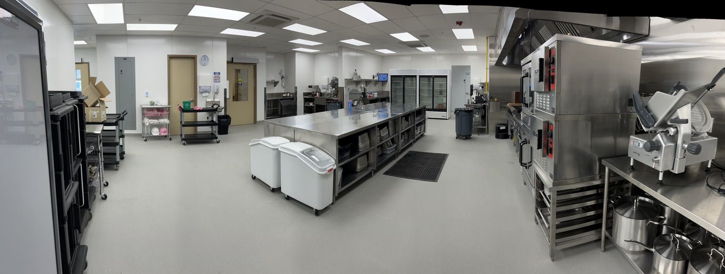 Large clean commercial healthcare kitchen with stainless steel central island, ovens, appliances and refrigerators — smooth light gray seamless floor