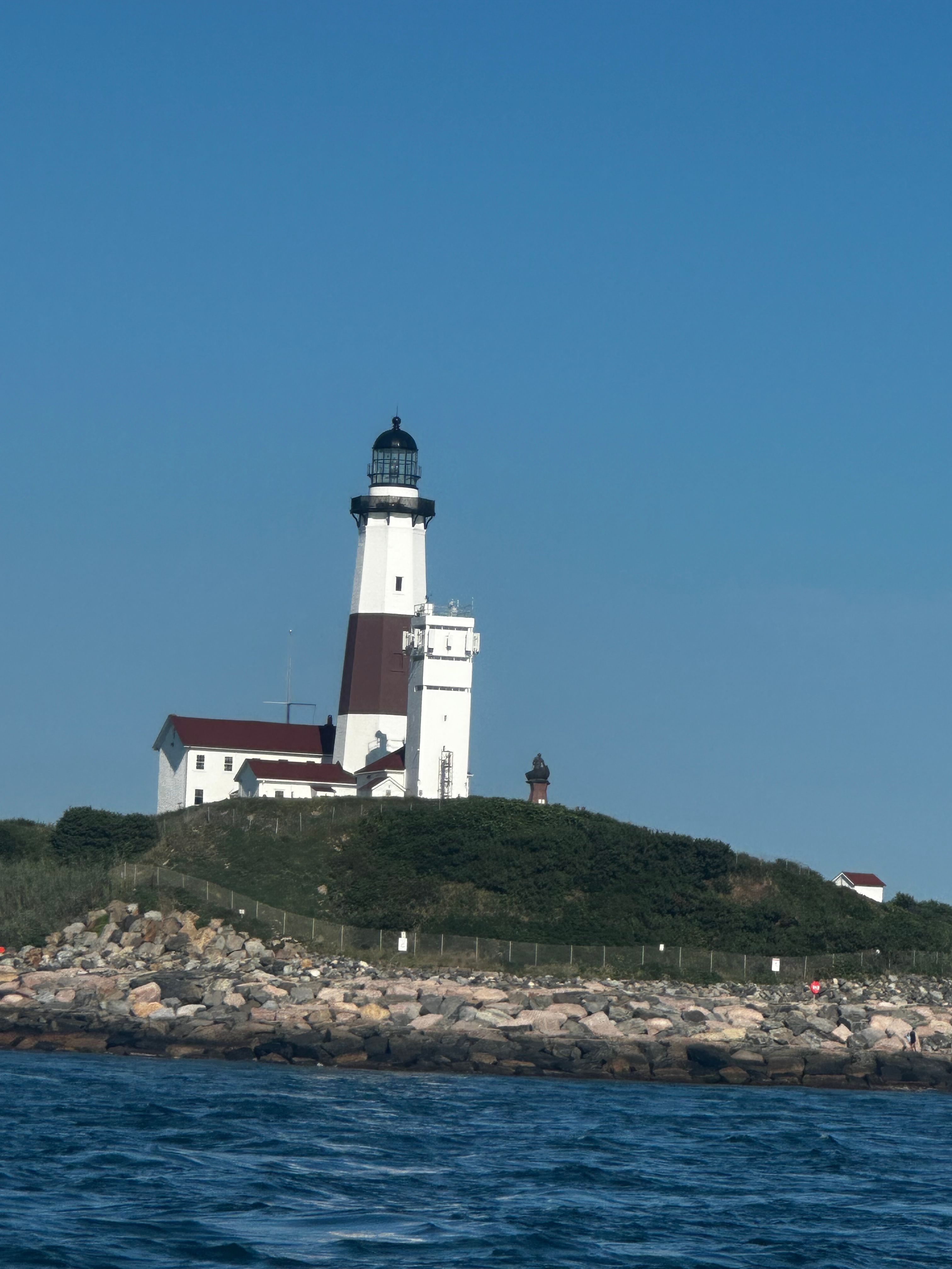Montauk Point Lighthouse, white lighthouse with dark band and black top on rocky shoreline, clear blue sky