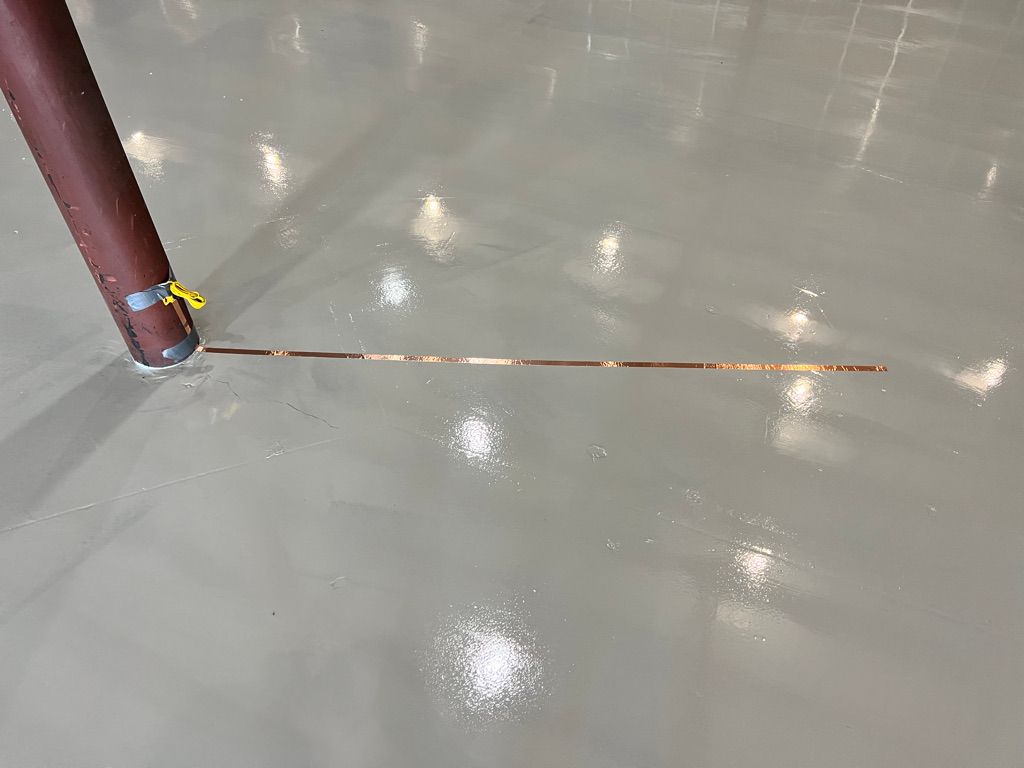 Shiny smooth gray ESD floor with copper grounding strip extending horizontally from base of brown pole secured with yellow tie