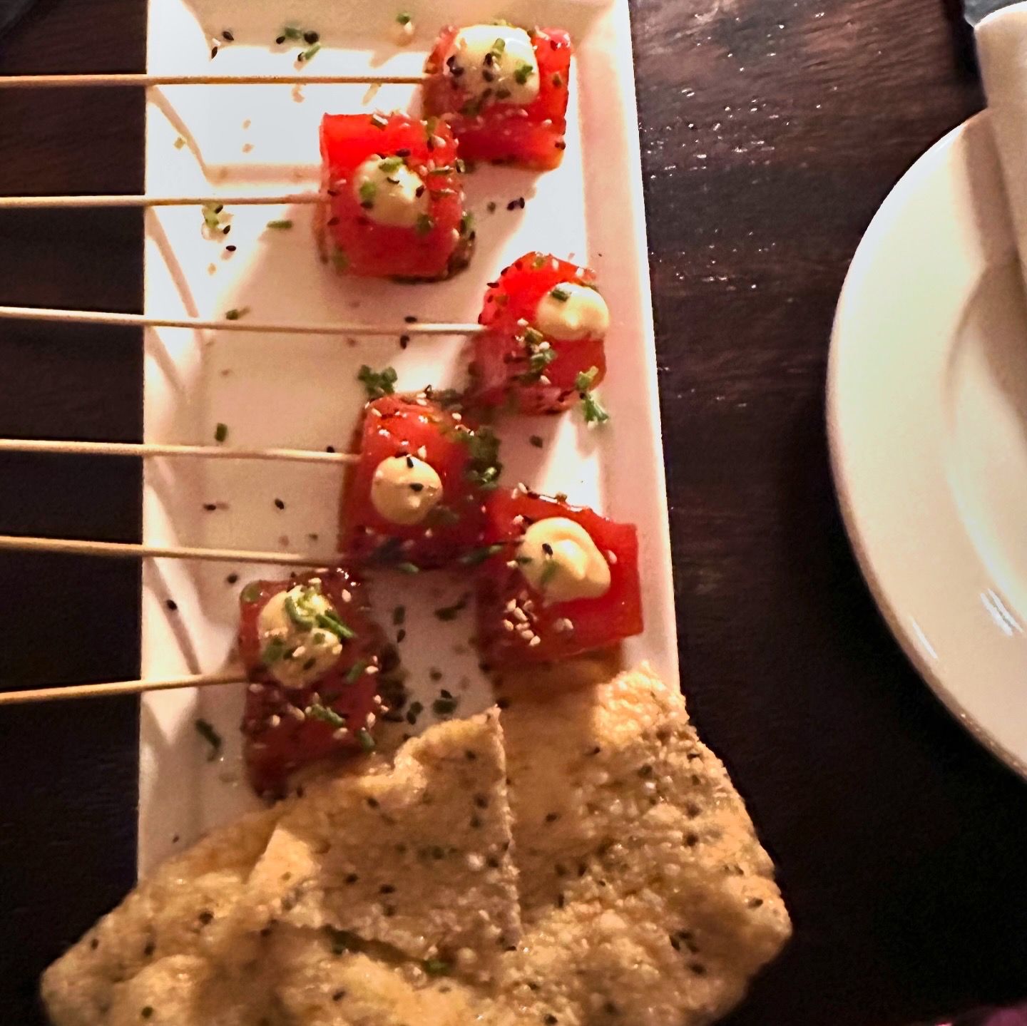 Santaella tuna tartare — vibrant red tuna cubes with cream dots, sesame seeds, chives, and crispy crackers on a white rectangular plate