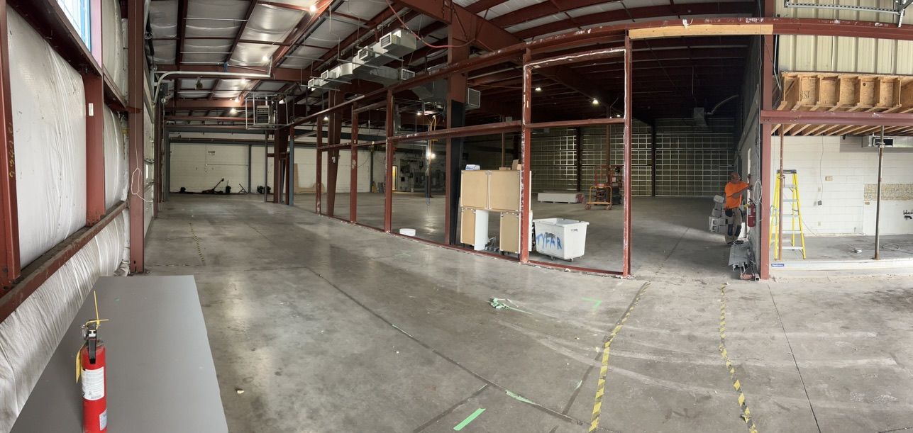 Large industrial warehouse with concrete floors, steel beams, fire extinguisher on table and worker in orange shirt preparing site for ESD flooring installation