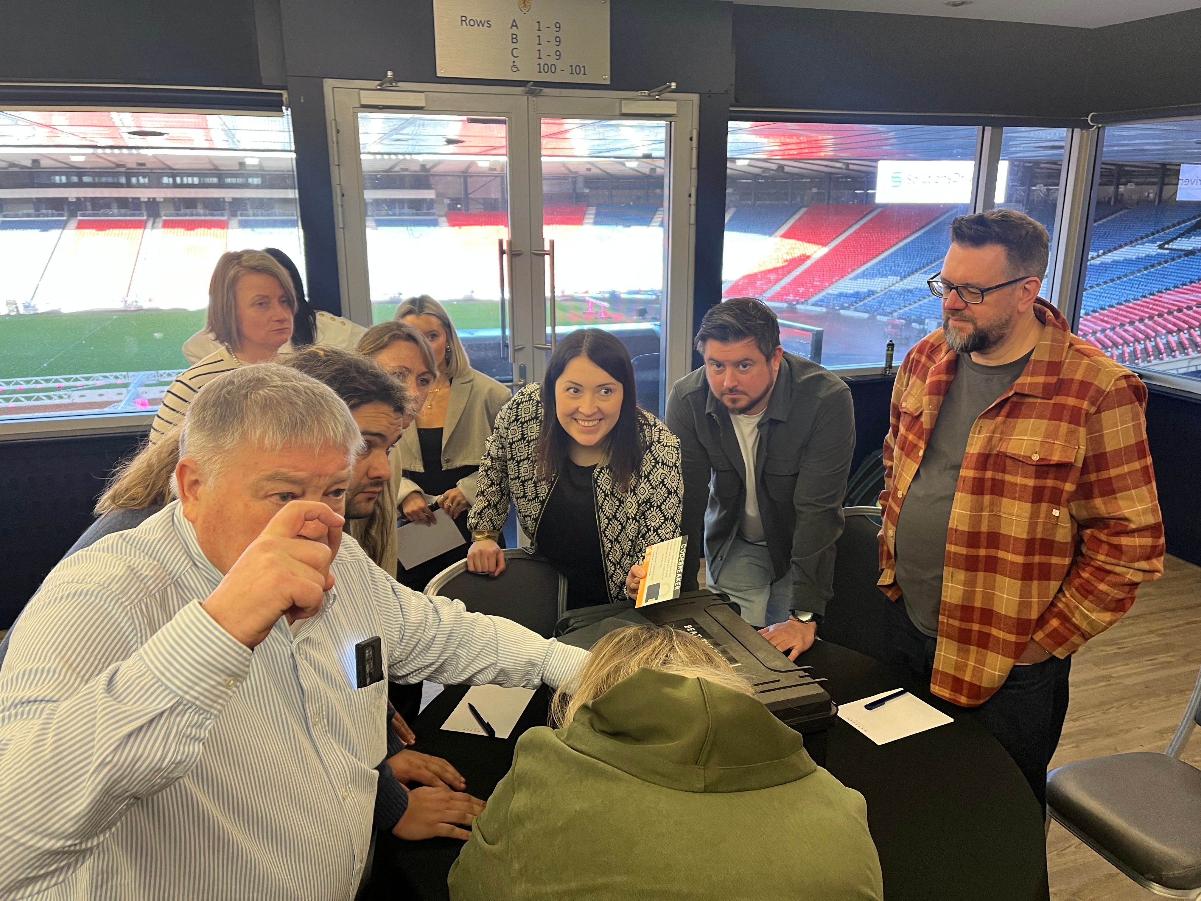 Beat the Box corporate team building — nine colleagues gathered around a table with the Beat the Box case, stadium seating visible through the windows